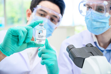 Couple male scientist wearing protection suit holding Coronavirus vaccine and working with many lab equipment for research vaccine at laboratory.