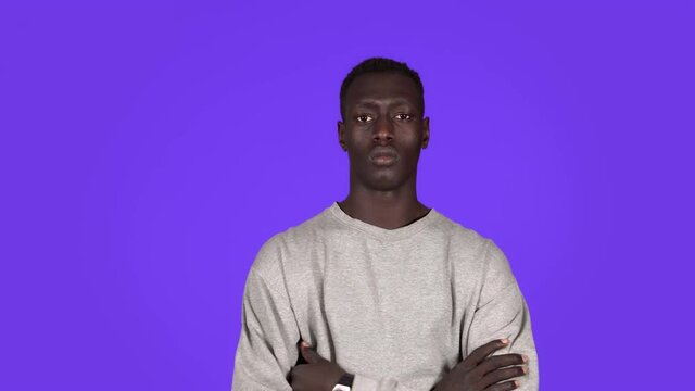 Portrait of young serious man staring at camera with crossed arms protesting. Beautiful african black man in white sweatshirt on blue background