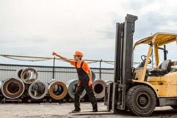 Young man builder in overalls and orange protective helmet near a construction loader machine on the construction metal coil store