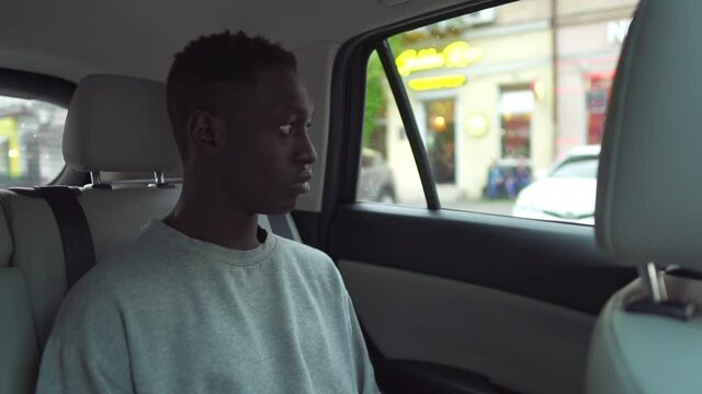Handsome young black man rides on back seat of a car, looks around in wonder out of the window. Big city view reflected in window. Wearing white casual shirt. Slow motion