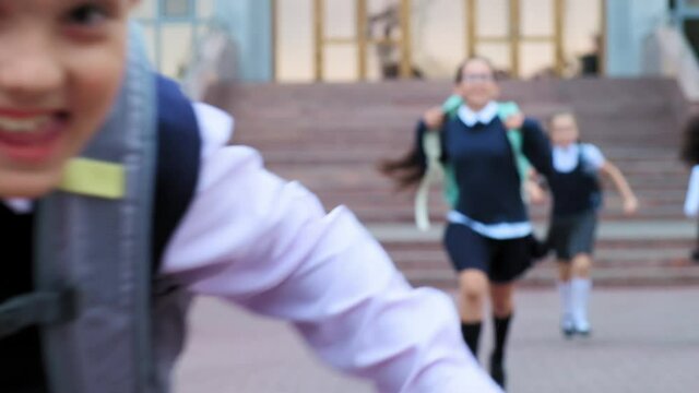 positive junior schoolboy in uniform with backpack and friends run along spacious school yard on warm summer day slow motion