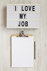 'I love my job' on a lightbox, clipboard with blank sheet of paper on a white wooden background, top view. Flat lay, from above, overhead.