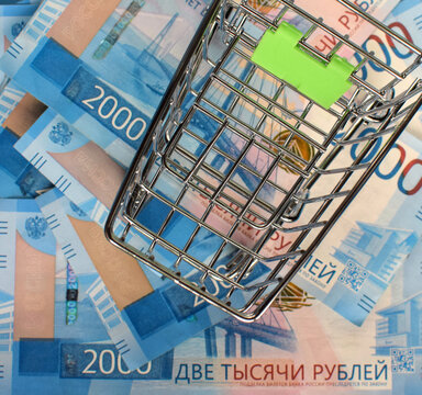 Russian Rubles Under The Basket On A Blue Background. Empty Grocery Cart On The Background Of 2000 Ruble Bills. View From Above. Selective Focus.