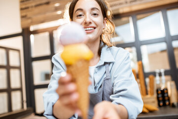 Female cheerful seller holding yummy ice cream in waffle cone indoors, selling sweets in the shop