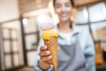 Female cheerful seller holding yummy ice cream in waffle cone indoors, selling sweets in the shop