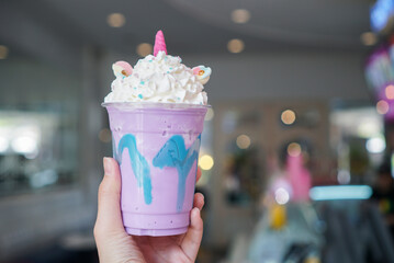 Unicorn Frappe. Hand holding a cup of unicorn smoothie, rainbow ice cream mixed with blueberry syrup topped with whipping cream, white chocolate and marshmallow. (Blurred background) © Swanya