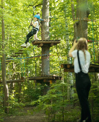 A woman taking a photo of her little daughter hanging on the insurance belt in the forest