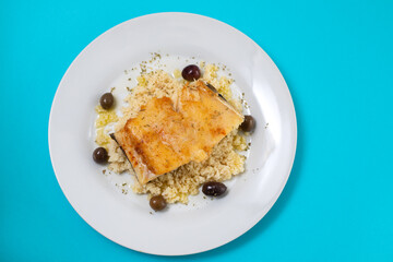 fried cod fish with cous cous and olives on white plate on blue background