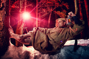 Medieval monk in canvas sackcloth robe with lattern resting on snow in dark forest and red light on...