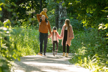 Obraz premium Young family of parents and two little kids walking in the forest - mom and dad holding their daughter on skate rollers by the hands