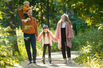 Fototapeta premium Young happy family of parents and two kids walking in the park - mom and dad holding their daughter on skate rollers by the hands