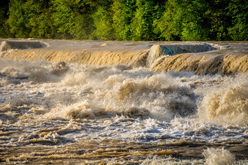 Raging flooded river flowing over man made cascades. Brown dirty muddy water flowing downstream violently during flooding. Dangerous situation after heavy rainfall in spring season