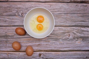 In selective focus top view of a fresh egg in a white bowl with brown eggs on old wooden table background 