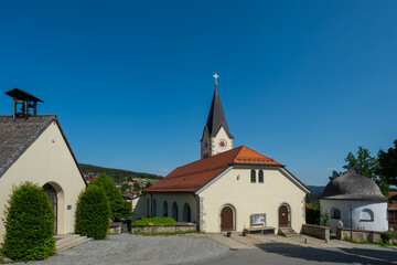 Fototapeta na wymiar Pfarrkirche und Kirchplatz im Ortskern von Sankt Englmar im Bayerischen Wald in der Oberpfalz in Bayern im Sommer bei blauem Himmel