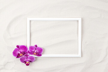 Top view of white photo frame on sand background decorated with purple orchid flowers. Summer and...