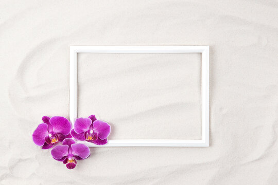 Top view of white photo frame on sand background decorated with purple orchid flowers. Summer and vacation concept. Flat lay. Copy space.