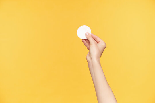 Cropped shot of young pretty woman's hand holding white cotton pad with fingers while being isolated over orange background. Human hands and beauty concept