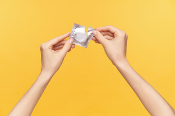 Cropped shot of young raised female's hands keeping silver pack with condom and unpacking it while being isolated over orange background. Relations and sex concept