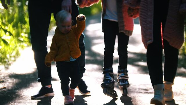 Young family of parents and two little kids walking in the summer park - little baby walking and a girl skating on the rollers