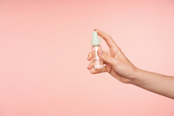 Studio photo of young female's fair-skinned hand keeping forefinger on button of bottle while going to spray content, isolated over pink background