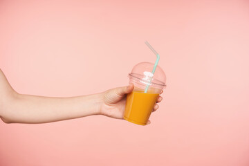 Indoor shot of raised pretty female's hand with nude manicure offering someone orange juice and reaching out ahead, isolated against pink background