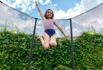 A girl jumping on a trampoline under the blue sky with clouds and the green background of ivy 
