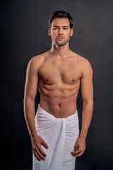 Fototapeta premium Handsome young bearded man isolated. Topless muscular man in towel is standing on grey background. Serious man looking at camera.