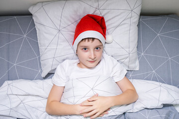 A preteen boy lying in bed in santa hat during christmas eve night and waiting for gifts and miracles, happy xmas and new year holiday