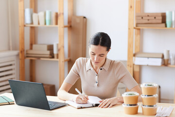 Serious woman sitting at the table and concentrating on her work she making notes in note pad and registering food delivery