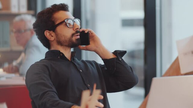 Young Handsome Male Architect Of Middle Eastern Ethnicity Talking On Mobile Phone While Sitting At Desk In Open Space Office