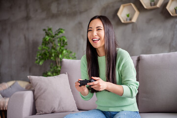 Photo of nice content chinese girl play video game hold joystick enjoy speed race laughing sit divan in house indoors