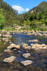 Fototapeta na wymiar The rocky Ohinemuri River in the Karangahake Gorge, New Zealand, surrounded by wooded hills. A suspension bridge crosses the river in the background