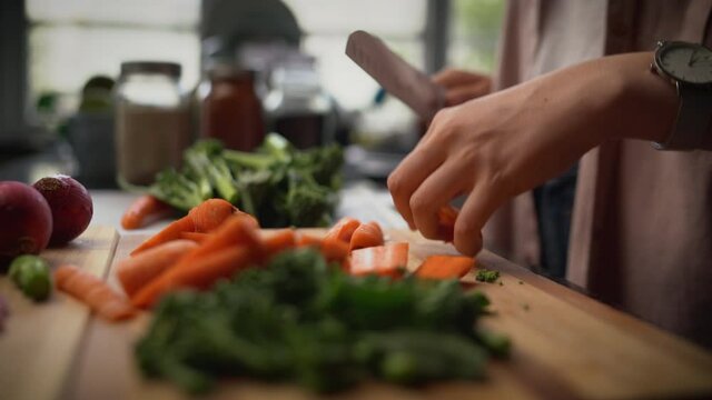 Close up of a woman slicing carrot with kitchen knife on cutting board and preparing salad in the kitchen