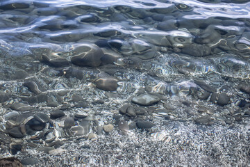 Closeup of transparent clear calm water surface with small grey pebbles sea bottom texture with waves optical distortion and caustics light effects. Trendy neutral abstract nature background