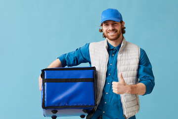 Portrait of young man working in delivery service he smiling at camera and showing thumb up against the blue background