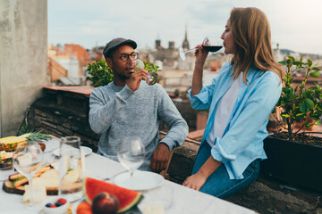 Happy young couple in love in casual clothes drinking wine dating on the restaurant rooftop with beautiful view on Barcelona, interracial young people having dinner at cafe terrace with healthy food