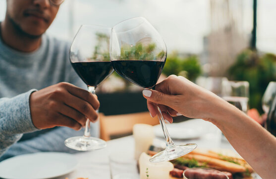 Young Happy Couple Having Date Dinner Celebrating Outdoors Making Cheers With Red Wine Glasses, People Enjoying Food Drink , Multiracial Couple Having Romantic Dinner At Outdoor Restaurant Terrace