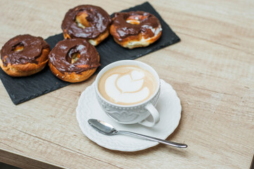 Coffee with a drawn heart and milk on a wooden table in a coffee shop. Chocolate donut with scattering on the table next to the coffee.