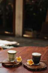 Vertical photo of cup of matcha latte green tea, glass of coffee with milk, yellow flowers, spoons on wooden boards on table