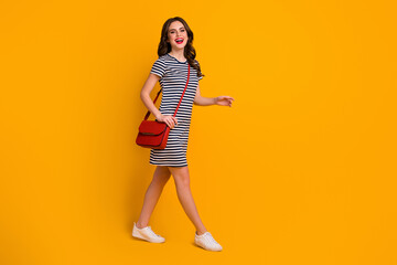 Fototapeta na wymiar Full body profile photo of pretty millennial lady red lips walk street sunny weather day shopping weekend wear casual striped short dress clutch footwear isolated yellow color background