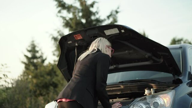 Baffled blonde comes, looks under hood of broken car and confusedly looks around