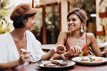 Portrait of charming tanned women in good mood eating tasty food in street cafe. Attractive brunette girls gossip and smile outside