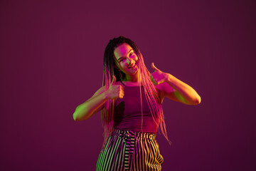 Young caucasian woman showing thumbs up on pink studio background in neon light. Beautiful model with dreadlocks. Human emotions, facial expression, sales, ad concept. Freak's culture. Copyspace.