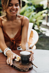 Pretty tanned woman in bra smiles sincerely and holds cup of coffee with milk. Blurred effect. Photo of girl in brown top posing near wooden table with glass of latte