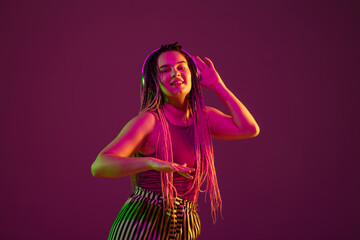 Young caucasian woman dancing with headphones on pink studio background in neon light. Beautiful model with dreadlocks. Human emotions, facial expression, sales, ad concept. Freak's culture.