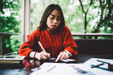 Concentrated female student in sweater doing homework task making creative drawings on terrace, talented graphic designer making sketches for project using stationery and equipment on working place