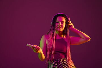 Vlog, selfie. Young caucasian woman with headphones, phone on pink studio background in neon. Beautiful model with dreadlocks. Human emotions, facial expression, sales, ad concept. Freak's culture.