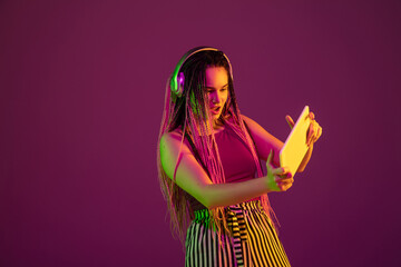 Playing. Young caucasian woman with headphones, tablet on pink studio background in neon light. Beautiful model with dreadlocks. Human emotions, facial expression, sales, ad concept. Freak's culture.
