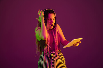 Shocked with headphones, tablet. Young caucasian woman on pink studio background in neon light. Beautiful model with dreadlocks. Human emotions, facial expression, sales, ad concept. Freak's culture.
