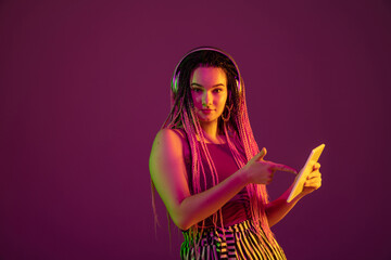 Pointing. Young caucasian woman with headphones, tablet on pink studio background in neon light. Beautiful model with dreadlocks. Human emotions, facial expression, sales, ad concept. Freak's culture.